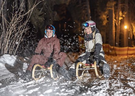 Zwei Personen rodeln nachts lachend auf Holzschlitten eine verschneite Piste hinunter, umgeben von wirbelndem Schnee und warmem Laternenlicht.