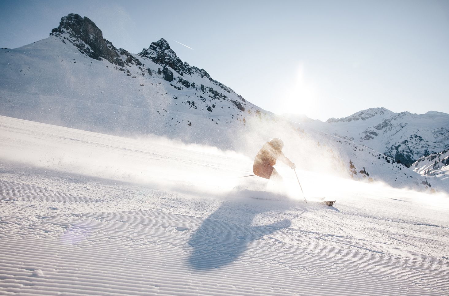 The image shows a skier in a fast-paced descent, surrounded by an impressive mountain landscape. The low-standing sun and the swirling snow add dynamism and a touch of winter magic to the scene.